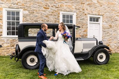 A joyful bride and groom share a laugh as she steps out of a vintage cream-and-black car in front of a historic stone building in Lancaster. The groom, dressed in a navy suit and brown shoes, helps the bride with her full lace gown as she holds a colorful bouquet of blue, pink, and purple flowers and wears vibrant blue shoes.