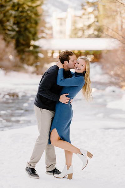 A couple sharing a quiet moment by the river during their winter engagement session in Vail, Colorado. The frozen river and snowy forest reflect the winter magic of the mountains, while their laughter and love create the perfect scene of authenticity and romance