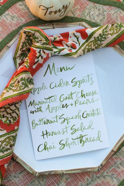 Menu card with green lettering and white mini pumpkin place card with gold calligraphy for fall table.