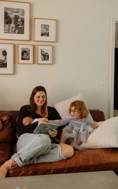 A mother holds her newborn in her lap while reading to her toddler. The mother and children sit on the couch as fine art New England Photographer takes beautiful candid photos. 