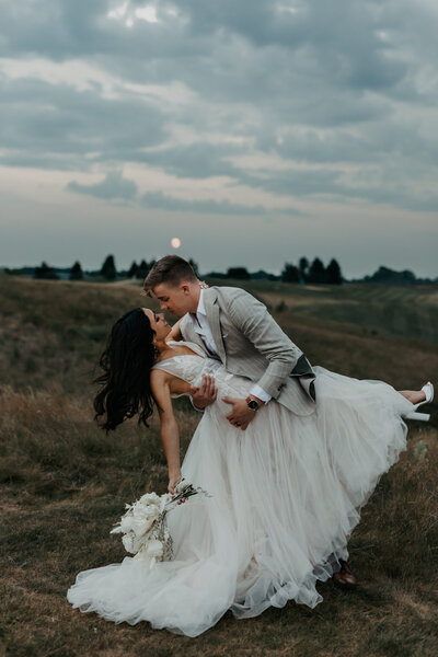 bride and groom dip kiss under the full moon overlooking a field of grass