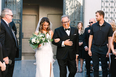 A bride walks down the aisle arm-in-arm with her father at SoHo63, a Chandler wedding and event venue. She holds a lush white and green bouquet and smiles softly, while her father beams with emotion as guests look on.