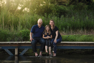 Family of 3 Mom, dad, and daughter sitting on a dock splashing water with green trees in the background