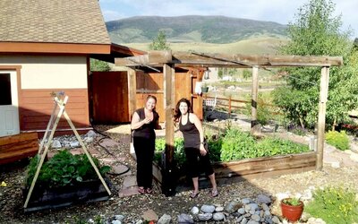 Two brunette women stand in front of a green, overflowing, garden bed that they planted. 