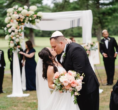 Whimsical floral wedding arch with colorful blooms and draping greenery by Leigh Florist, framing the couple's ceremony.