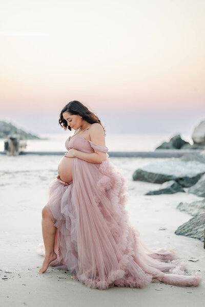 An expectant mother in a pink tulle gown cradles her belly. She is posed on the beach at sunset.