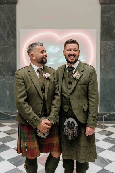 A wedding portrait of an LGBTQ+ couple in Aberdeen Art Gallery following their wedding. The stand in front of a large neon love heart.