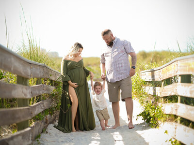 A family of three walks on a sandy beach path. The pregnant woman in a green dress holds her son's hand with the man on the other side. The mood is joyful.