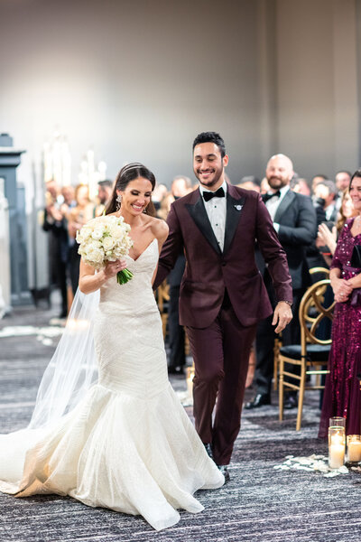 A smiling bride in a white wedding dress and a groom in a dark maroon tuxedo walk down the aisle together, hand in hand. They are surrounded by applauding guests seated at elegant chairs, with candles and soft lighting creating a warm ambiance.