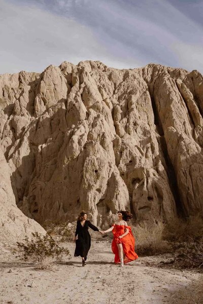 A couple shares a moment during their elopement, standing on a fallen tree in a golden meadow surrounded by tall pines, with a small rustic chapel and granite cliffs in the background in Yosemite National Park.