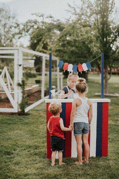 A girl stands behind the counter of a play popsicle stand as a boy holding a girls hand waits in front