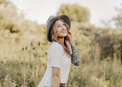 woman in white t-shirt, hand on brim of grey hat looking up toward the tree with field in background