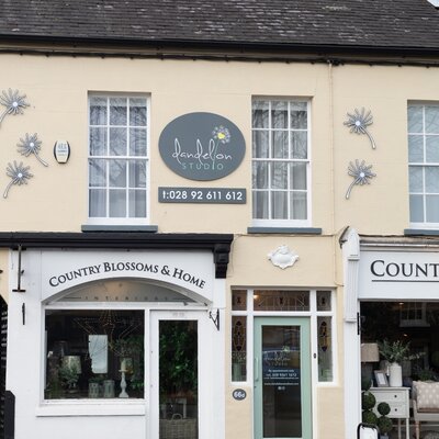 The exterior of Dandelion Studio in Moira, Northern Ireland. The storefront features a sign with the studio’s name, logo, and contact number displayed above the door. The entrance, marked by a soft green door with glass panels, sits between two large windows. On the left side, there’s a neighbouring shop named "Country Blossoms & Home." The studio’s branding includes decorative dandelion motifs on the building, creating a charming and inviting appearance.