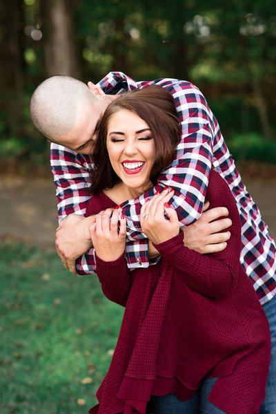 Guy surprises his fiance with a hug and kiss during their engagement photos, she's wearing a maroon top and bright red lipstick and he's in a checkered shirt