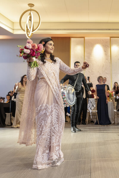 An Indian woman in a sparkling traditional outfit dances while holding a vibrant bouquet of flowers. A musician follows her playing a drum. Guests in formal attire watch and smile in the background. The setting appears to be an indoor celebration or event.