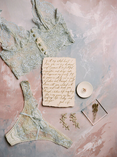 Heirloom bridal lingerie from Claire Pettibone with soft blue lace beside hand calligraphed vows on an abstract painted surface