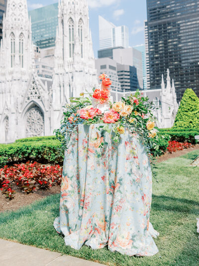 wedding cake sits atop a floral linen covered table with matching florals around it with St. Patrick's Cathedral in the background in New York City