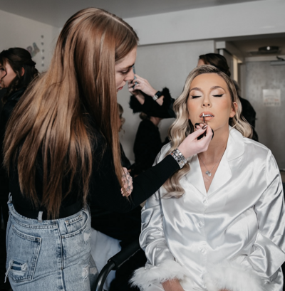 Makall applying lipstick to bride in a white silk robe on wedding morning