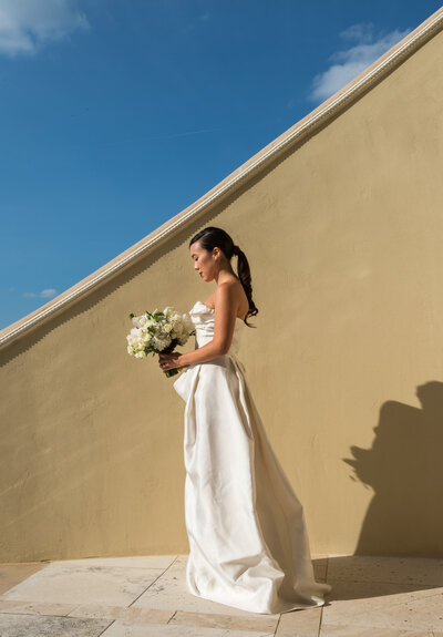 Bride standing gracefully in front of a vibrant yellow stucco wall with a blue sky above, capturing a stunning contrast in her wedding attire.
