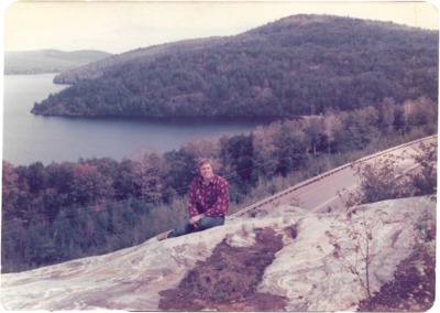 a man sitting on a rock with a treed mountain and lake behind him.