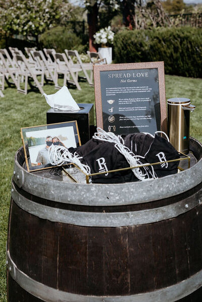 Spring wedding safety setup at the Palm Event Center in Pleasanton, CA, featuring custom gold, black, and white face masks, a Covid-19 restrictions sign, and other sanitary items elegantly displayed on a wine barrel.