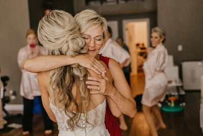 A mother, facing the camera, embraces her daughter right after she has put on her wedding dress.