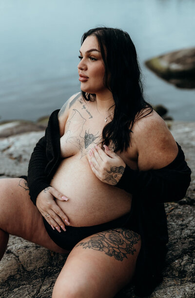 En gravid kvinna sittandes på ett berg vid vattnet med ena handen på sin gravidmage och andra över sitt bröst tittandes ut över vattnet