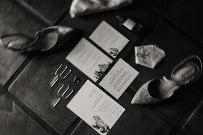 A black-and-white artistic flat lay of wedding details from a Tanque Verde Ranch wedding in Tucson, Arizona. The image includes wedding invitations, rings, and shoes, arranged on a textured surface.