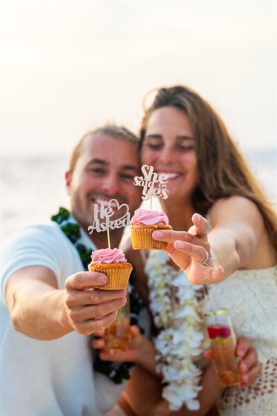 Engaged couple smiling and holding up decorated proposal cupcakes with He Asked and She Said Yes toppers during their sailboat celebration in Hawaii.