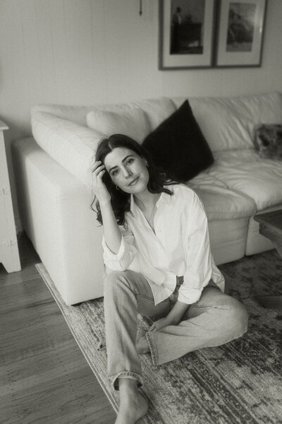 Woman wearing white button down and jeans sitting on a rug in front of a couch looking at camera and pushing hair out of face