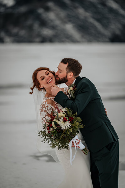 Groom gives bride lots of kisses in the snow making her giggle.