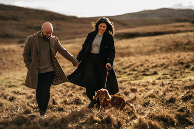 Couple holding hands and walking their happy dog through a sunlit field during golden hour in the countryside.