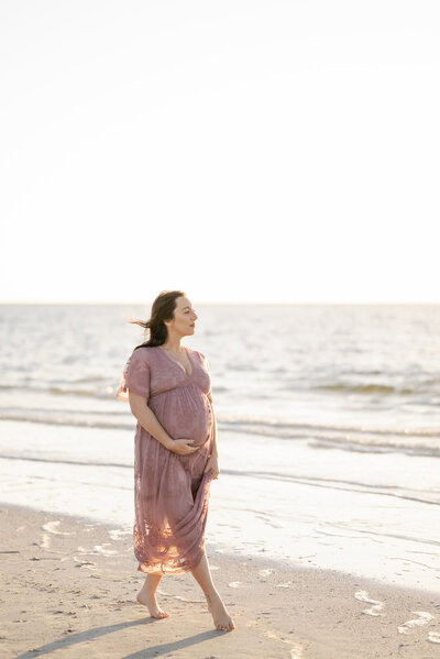 Pregnant woman walking along the beach near the water's edge looking out toward the setting sun