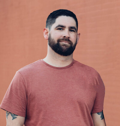 A man with short dark hair and a mustache, wearing a black button-up shirt over a white T-shirt, stands against a plain background, smiling as if pleased with his latest web design and branding project.