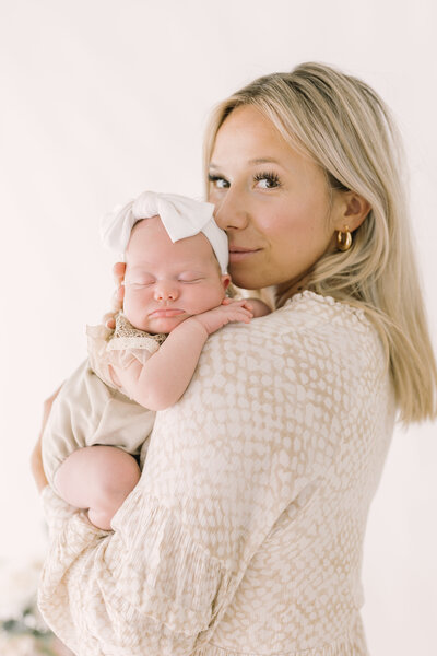 A mother holding her newborn baby close in a natural light studio, both wrapped in soft neutral tones — Raleigh portrait photographer.