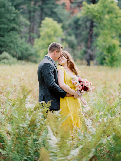 Sedona Engagement Session Photographer Couple is cuddling in a field holding a pink bouquet wearing a yellow dress