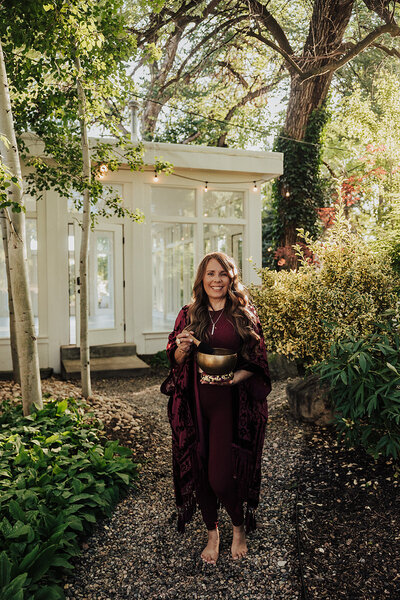Patty in a maroon dress with her gold sound bowl on a path  with trees