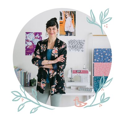 A portrait of artist and designer Skye McNeill in her studio. Image shows Skye McNeill in a bold printed blazer, her arms crossed gently. She stands in front of a table with a sewing machine, pantone books, and a selection of textile design books. Next to her are fabric samples of her textile designs and on the wall are images of her art and kids clothing design.