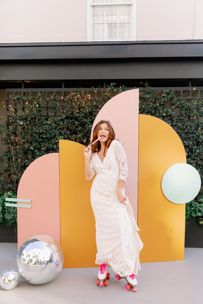 A woman in a white lace dress and pink shoes poses in front of a custom 'Pink Deco' backdrop by Shindig Social. The backdrop features pink and yellow geometric panels with reflective disco balls and a green sphere, creating a vibrant and stylish setting.