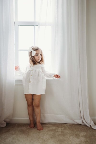 A young child in a white outfit standing by a window with sheer curtains during a Pittsburgh family photography session.