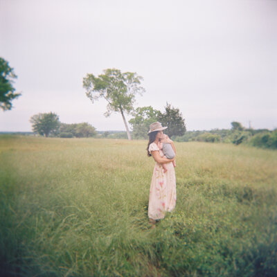 Film photo of mom in a long dress and hat holding her baby in a grassy field