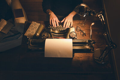 A close aerial view of a man typing on an antique typewriter. The room is dark save for the desk lamp that lights his space, which also features a journal and a pair of glasses.