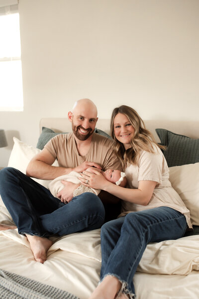 A mother and father sitting on their bed holding their newborn baby girl.