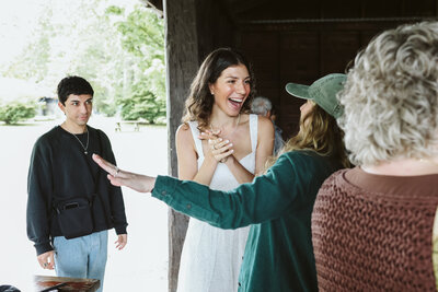 A joyful bride in a white dress claps and smiles while engaging with guests at an outdoor wedding reception. A woman in a green hat gestures animatedly, while others, including a young man, look on.
