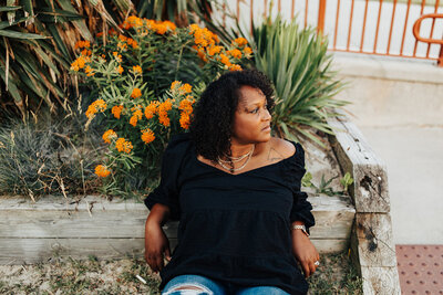 Overhead shot of black female leaning against a garden bed with a black off the shoulder shirt on and jeans.