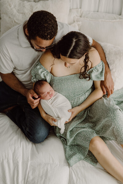 Newborn photo looking down at mom and dad sitting next to each other on a bed looking at newborn baby boy in white in Annapolis Maryland.