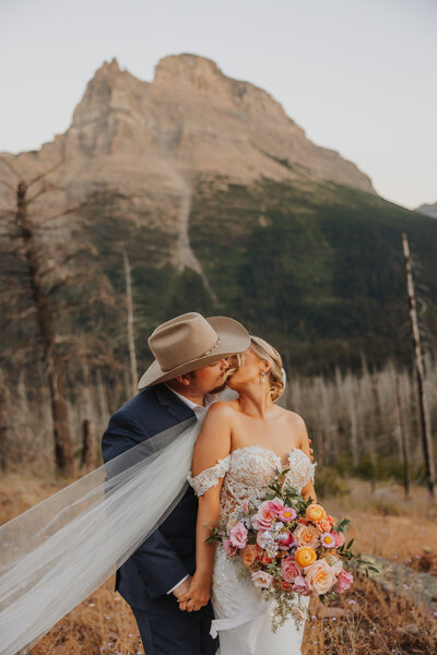 Groom in a cowboy hat kissing the bride while her veil blows in the wind.
