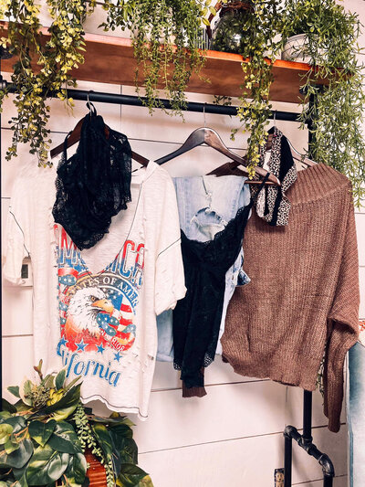 Curated outfits including a graphic tee, sweater, ripped jeans, and lacy underwear all hung on hangers and hung  surrounded by draping greenery