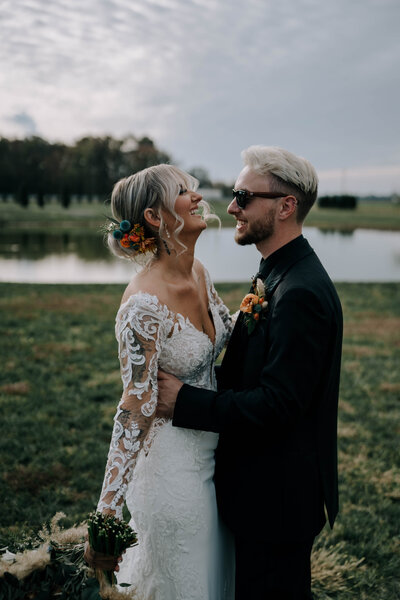 Bride with an elegant updo and soft, wispy face-framing pieces smiles at her groom by the water on their Maryland wedding day — hair by Klover Beauty.
