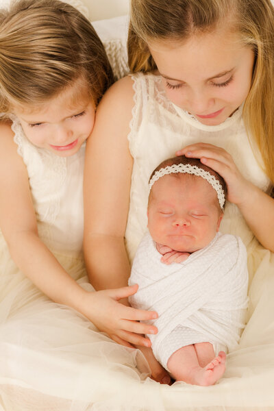 Two sisters hold newborn wearing white dresses.